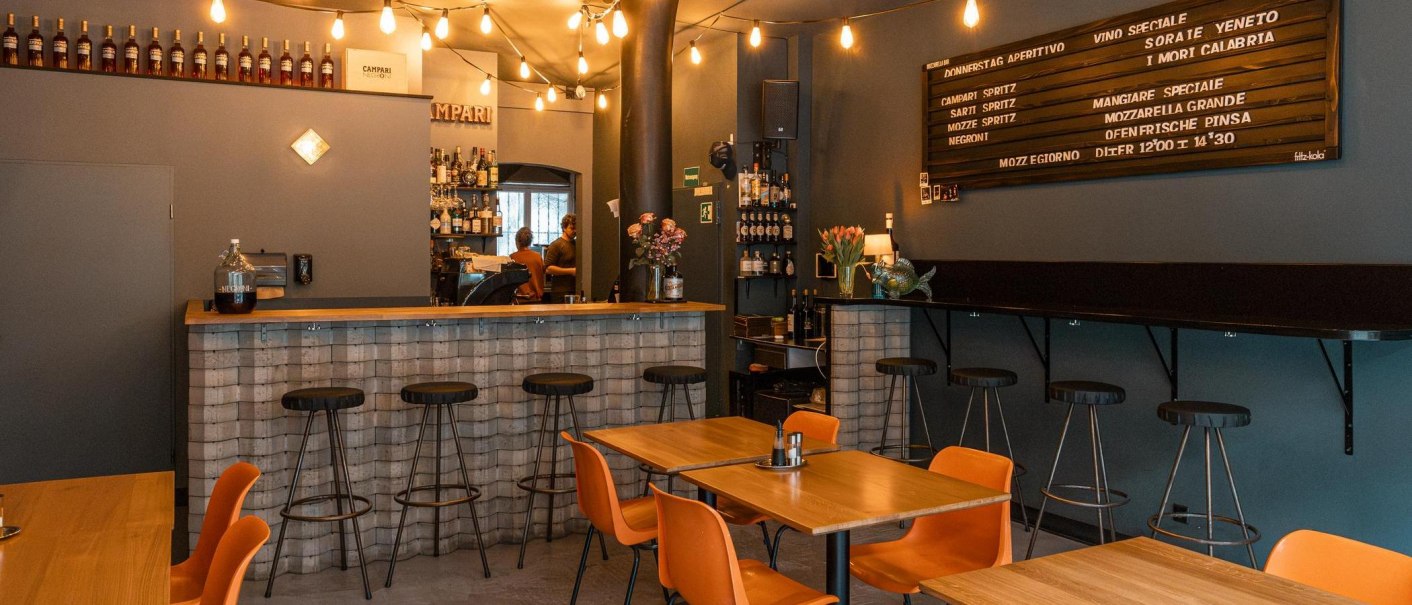 Gemütliche Bar mit Holztischen, Barhockern und einer Theke. Lichterketten und ein Menübrett schmücken die Wand., © SMG, Sarah Schmid Gemütliche Bar mit Holztischen, Barhockern und einer Theke. Lichterketten und ein Menübrett schmücken die Wand., © SMG, Sarah Schmid