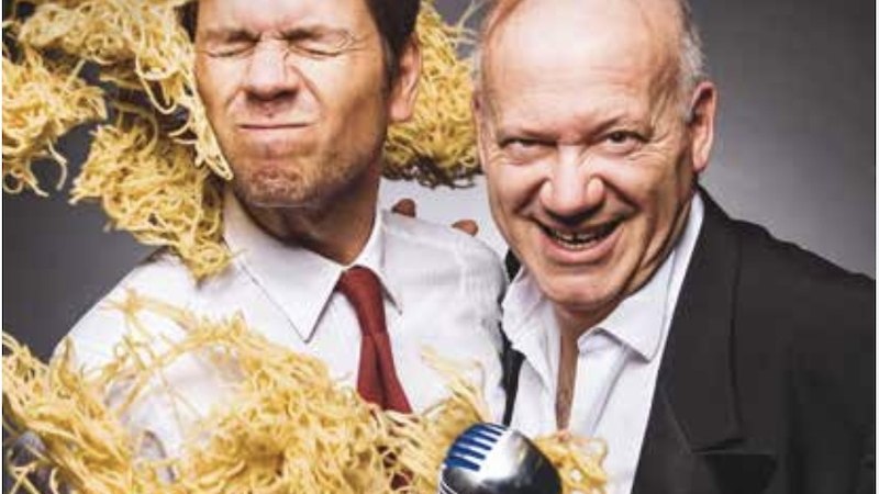 Zwei M&auml;nner in Anz&uuml;gen, einer mit Nudeln im Gesicht, der andere h&auml;lt ein Mikrofon. Beide l&auml;cheln und wirken humorvoll., &copy; Renitenztheater Stuttgart e.V.