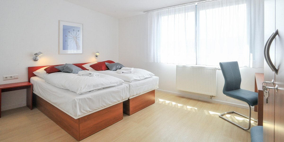 A bright double room with two single beds, red and gray pillows, a desk with a chair, and a large window with white curtains., &copy; G&auml;stehaus ABZ Stuttgart
