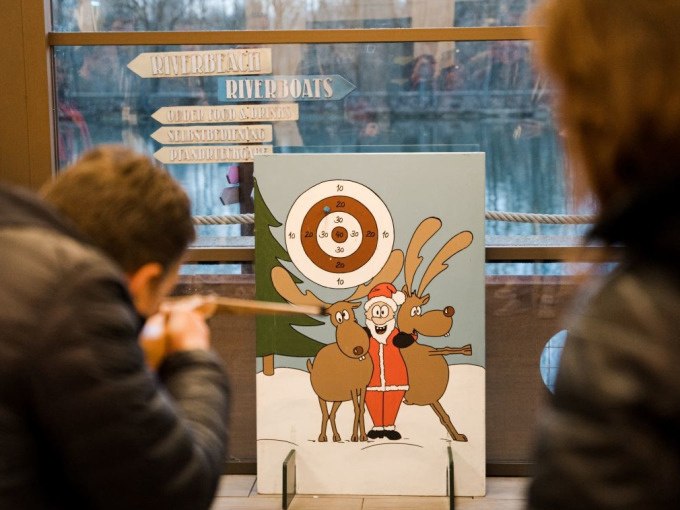 Zwei Personen zielen mit Blasrohren auf eine Zielscheibe mit einem Weihnachtsmann und Rentieren. Im Hintergrund sind Schilder und ein Fenster zu sehen., &copy; Cool-Tours StattReisen