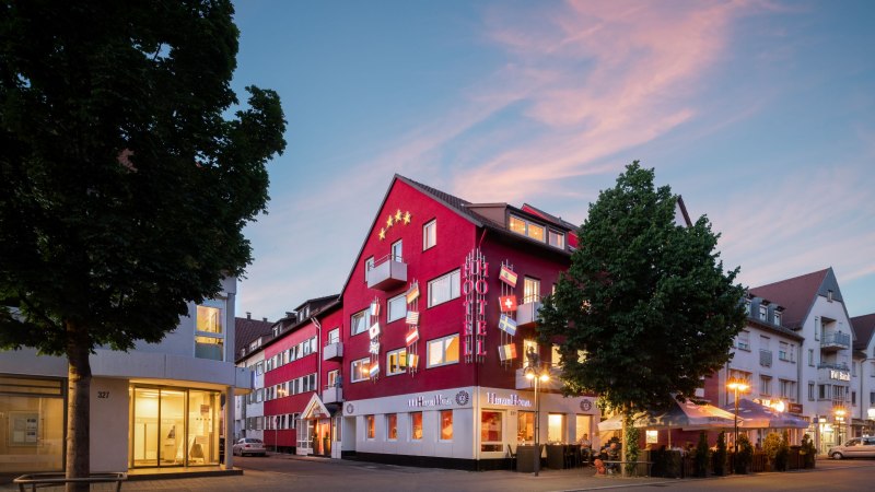 Rotes Hotelgebäude mit Flaggen und beleuchteten Schildern bei Dämmerung, umgeben von Bäumen und Straßenlaternen., © Hetzel Hotel Stuttgart Rotes Hotelgebäude mit Flaggen und beleuchteten Schildern bei Dämmerung, umgeben von Bäumen und Straßenlaternen., © Hetzel Hotel Stuttgart