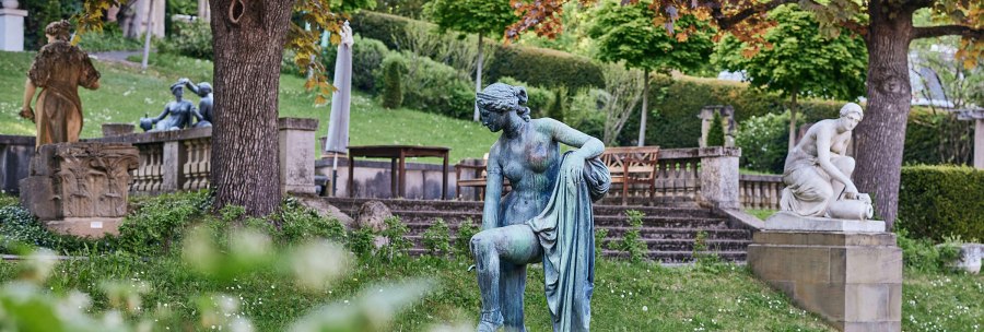Ein Garten mit mehreren Skulpturen, umgeben von Bäumen und Grünflächen. Im Vordergrund eine bronzene Figur, im Hintergrund weitere Statuen., © Städtisches Lapidarium, Foto Julia Ochs
