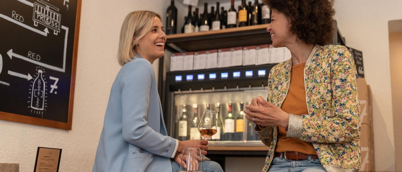 Two women are sitting on high stools, one of them is holding a glas of wine. They are laughing at each other. In the background is a wine dispenser with wine bottles that can be tapped., &copy; Stuttgart-Marketing GmbH, Martina Denker