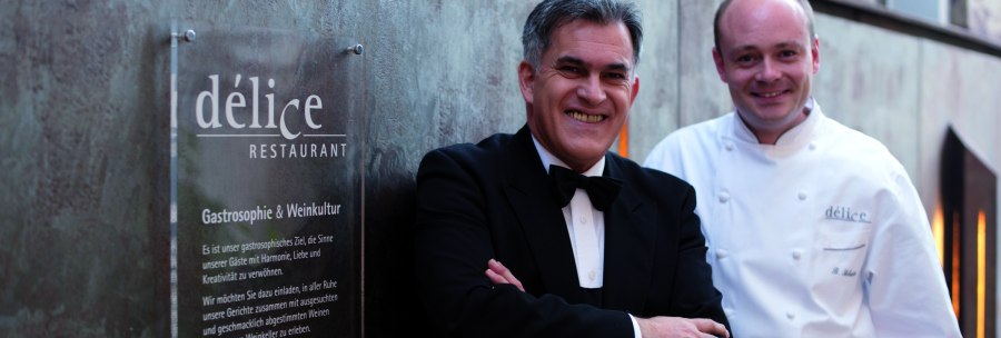 Two men stand in front of a sign for the D&eacute;lice restaurant. One is wearing a suit, the other a chef's jacket., &copy; Delice