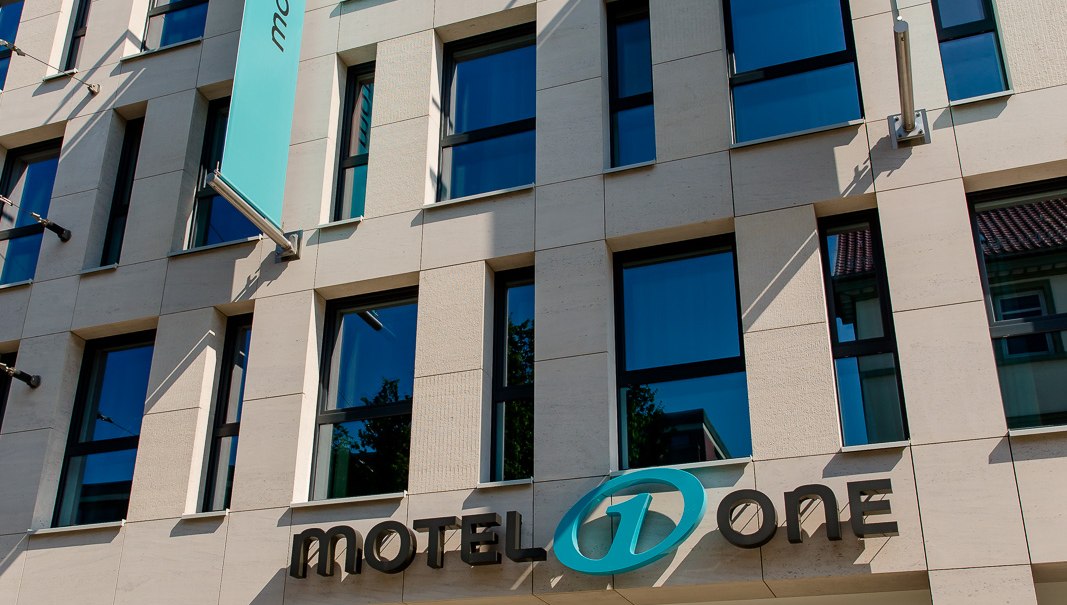 Modern fa&ccedil;ade of a building with Motel One logo and blue flag waving in the wind. Clear lines and large windows characterize the design., &copy; Motel One Stuttgart-Bad Cannstatt