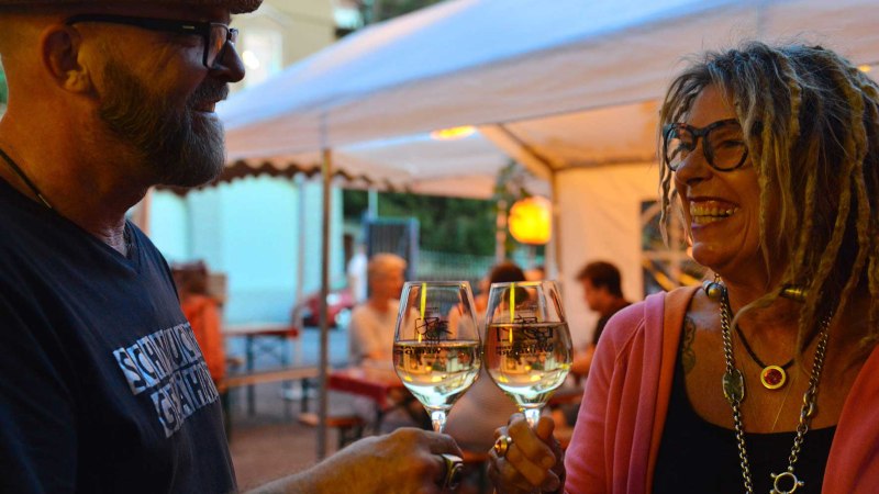 Zwei Personen sto&szlig;en l&auml;chelnd mit Weingl&auml;sern an, unter einem Zelt bei einem sommerlichen Event., &copy; Weingut Ruoff