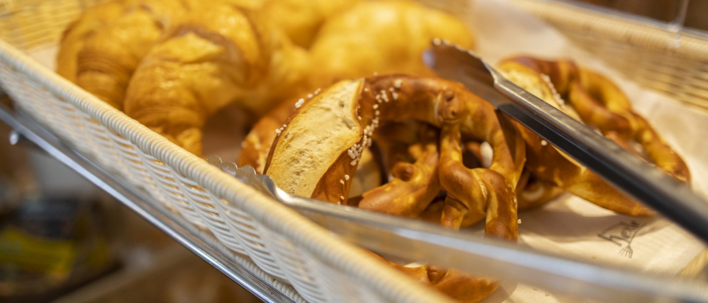 A basket of fresh croissants and pretzels, with tongs next to it for serving., © Hotel Gloria GmbH Restaurant Möhringer Hexle A basket of fresh croissants and pretzels, with tongs next to it for serving., © Hotel Gloria GmbH Restaurant Möhringer Hexle