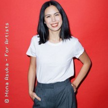 Eine Frau mit schwarzem Haar trägt ein weißes T-Shirt und graue Hose. Sie lächelt vor einem roten Hintergrund., © links im Bild