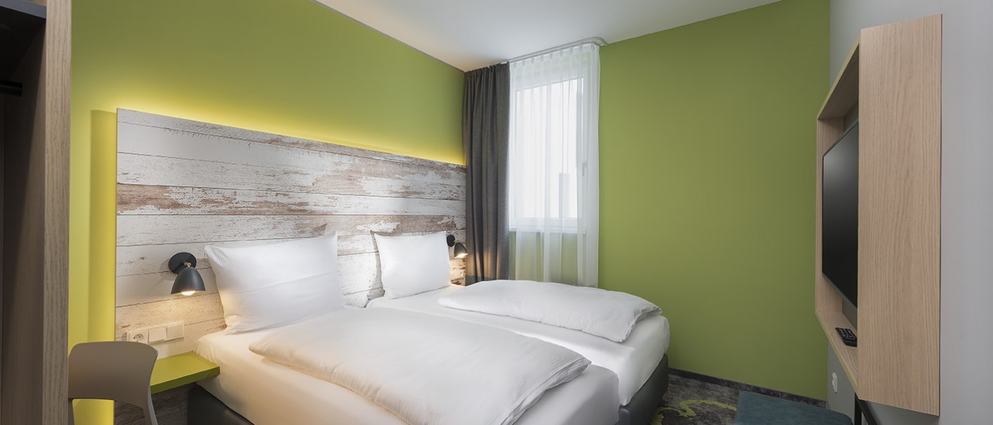 Modernes Hotelzimmer mit zwei Einzelbetten, grüne Wand, Holzkopfteil, Fernseher und Fenster mit Vorhängen., © ibis Styles Stuttgart Vaihingen Modernes Hotelzimmer mit zwei Einzelbetten, grüne Wand, Holzkopfteil, Fernseher und Fenster mit Vorhängen., © ibis Styles Stuttgart Vaihingen
