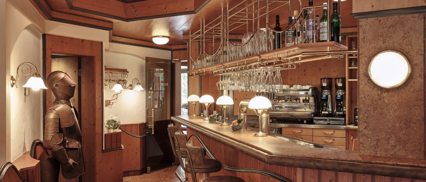 Gemütlicher Barbereich mit Holzverkleidung, Barhockern und einer Ritterrüstung. Gläser und Flaschen sind auf Regalen über der Theke platziert., © Badhotel Restaurant Stauferland