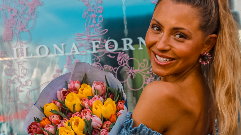 Frau mit Blumenstrauß lächelt vor einem Schaufenster mit dem Schriftzug 'Ilona Forner'., © Ilona Forner, Stuttgart