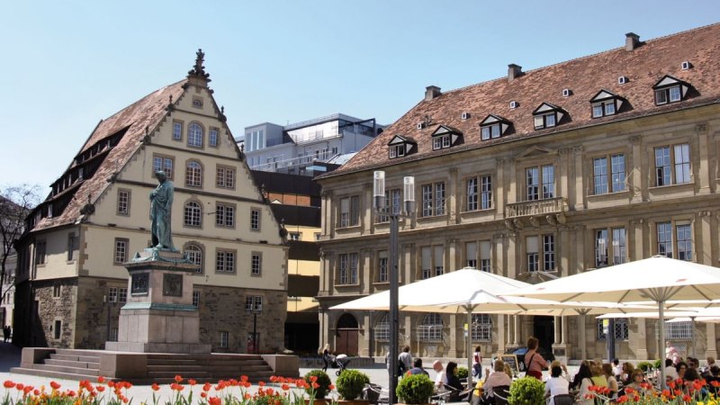 Schillerplatz in Stuttgart mit Fruchtkasten, Schillerdenkmal und Menschen in einem Café. Bunte Blumen im Vordergrund, blauer Himmel im Hintergrund., © Stuttgart-Marketing GmbH