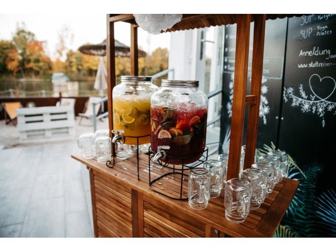 Drinks station with two large glasses full of colorful lemonade and many empty glasses on a wooden table, outdoors by a lake., © Cool-Tours StattReisen Drinks station with two large glasses full of colorful lemonade and many empty glasses on a wooden table, outdoors by a lake., © Cool-Tours StattReisen