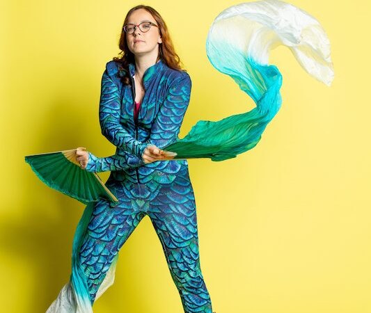Person in blau-grünem Kostüm mit Schuppenmuster und goldenen Stiefeln posiert vor gelbem Hintergrund, hält bunte Stoffbahnen in den Händen., © Rosenau Kultur e.V. Person in blau-grünem Kostüm mit Schuppenmuster und goldenen Stiefeln posiert vor gelbem Hintergrund, hält bunte Stoffbahnen in den Händen., © Rosenau Kultur e.V.