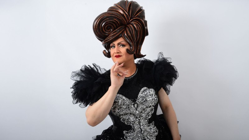 Person in auffälligem Kostüm mit großer Perücke und glitzerndem Kleid posiert vor weißem Hintergrund., © Friedrichsbau Varieté Person in auffälligem Kostüm mit großer Perücke und glitzerndem Kleid posiert vor weißem Hintergrund., © Friedrichsbau Varieté