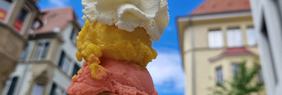 An ice cream cone with three scoops in different colors and whipped cream, against a background of buildings and blue sky., &copy; kesselglueck.de