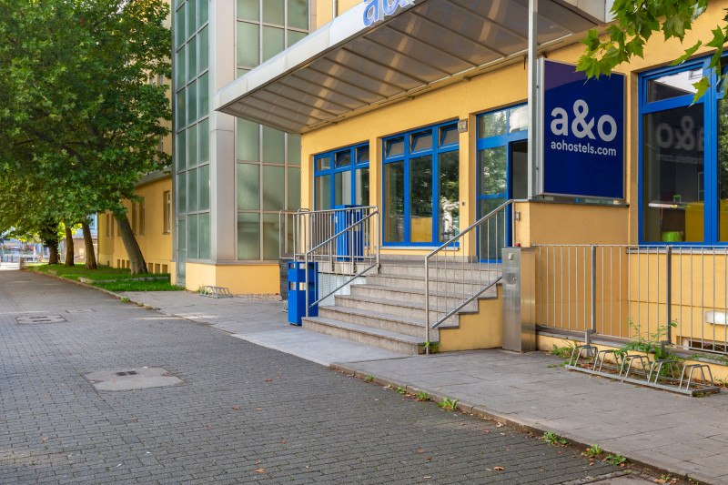 Yellow building with blue windows, stairs and canopy. Lettering 'a&o' and 'aohostels.com'. Tree and sidewalk in the foreground., © A&O Hostels Marketing GmbH Yellow building with blue windows, stairs and canopy. Lettering 'a&o' and 'aohostels.com'. Tree and sidewalk in the foreground., © A&O Hostels Marketing GmbH