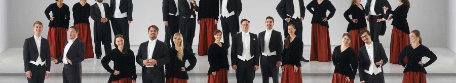 Der Kammerchor Stuttgart posiert in formeller Kleidung auf einer Bühne. Die Frauen tragen rote Röcke und schwarze Oberteile, die Männer Frack., © Jens Meisert