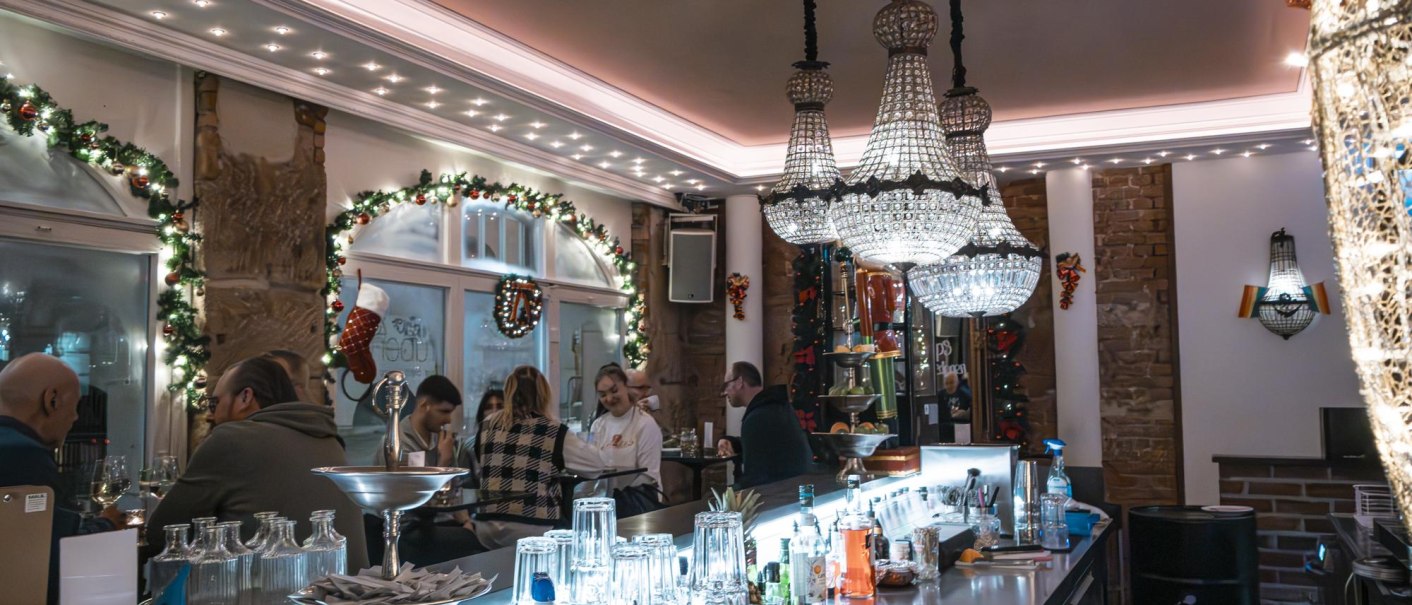 Festlich dekorierte Bar mit Kronleuchtern, Gästen an der Theke und weihnachtlichem Schmuck. Gläser und Flaschen stehen auf der Theke., © SMG Stuttgart Marketing GmbH - Sarah Schmid Festlich dekorierte Bar mit Kronleuchtern, Gästen an der Theke und weihnachtlichem Schmuck. Gläser und Flaschen stehen auf der Theke., © SMG Stuttgart Marketing GmbH - Sarah Schmid