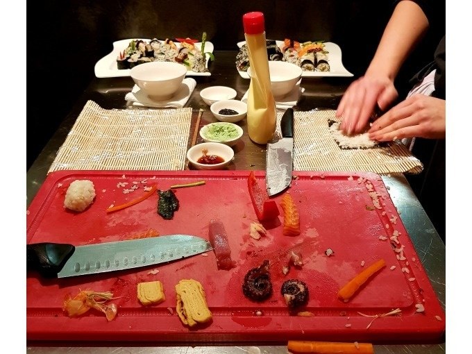 Sushi-Zubereitung mit Zutaten auf Schneidebrett, Bambusmatte und fertigen Sushirollen im Hintergrund., © Kultweine-Kochwelten-Kunstücke - Kochschule Sonja Lenz Sushi-Zubereitung mit Zutaten auf Schneidebrett, Bambusmatte und fertigen Sushirollen im Hintergrund., © Kultweine-Kochwelten-Kunstücke - Kochschule Sonja Lenz
