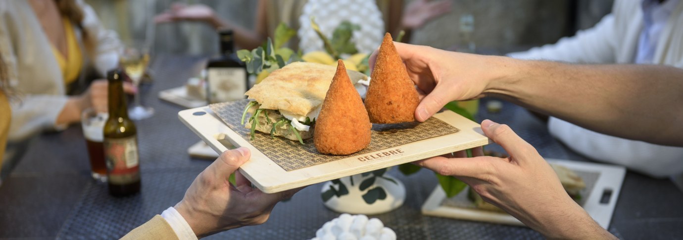 Zwei Personen reichen ein Holzbrett mit Arancini und einem Sandwich an einem Tisch mit mehreren G&auml;sten., &copy; CELEBRE