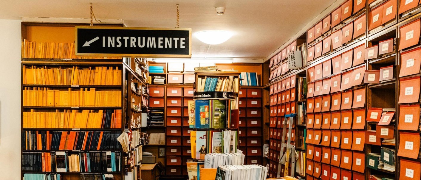 Ein Musikgeschäft mit Regalen voller Noten und einem Schild 'Instrumente'. Viele orangefarbene Ordner und Bücher sind zu sehen., © Stuttgart-Marketing GmbH, Sarah Schmid