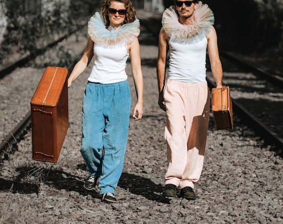 Zwei Personen mit Rüschenkragen und Sonnenbrillen gehen auf Bahngleisen. Sie tragen Koffer und lässige Kleidung in Blau und Rosa., © Rosenau Kultur e.V. Zwei Personen mit Rüschenkragen und Sonnenbrillen gehen auf Bahngleisen. Sie tragen Koffer und lässige Kleidung in Blau und Rosa., © Rosenau Kultur e.V.