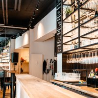 Moderne Bar mit hölzernem Tresen, Regalen voller Gläser und Flaschen. Helle Beleuchtung und stilvolle Einrichtung schaffen eine einladende Atmosphäre., © SMG_Sarah Schmid Moderne Bar mit hölzernem Tresen, Regalen voller Gläser und Flaschen. Helle Beleuchtung und stilvolle Einrichtung schaffen eine einladende Atmosphäre., © SMG_Sarah Schmid