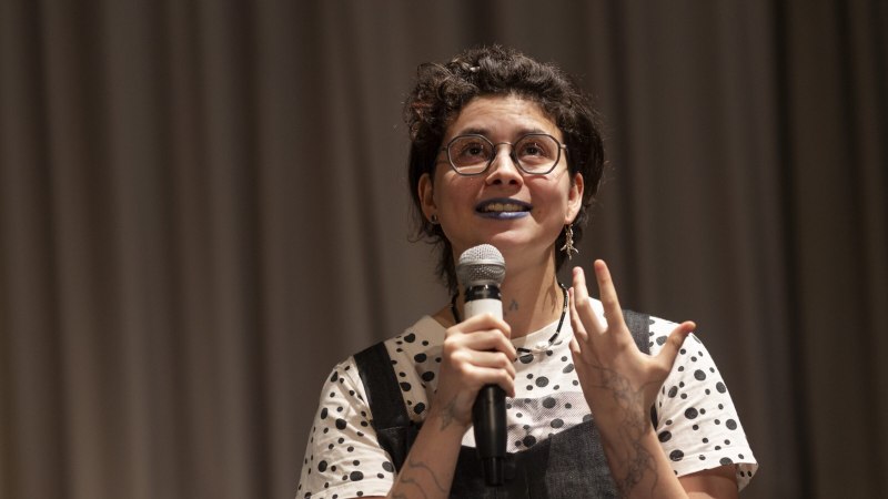 Eine Person mit Brille und blauem Lippenstift spricht in ein Mikrofon. Sie tr&auml;gt ein gepunktetes Oberteil und hat t&auml;towierte Arme., &copy; Astrid Ackermann