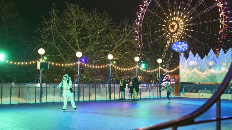 Eislaufbahn bei Nacht, beleuchtet mit Lichterketten. Menschen laufen Schlittschuh, im Hintergrund ein beleuchtetes Riesenrad und ein Schild 'Wintertraum'., © SMG, Sarah Schmid Eislaufbahn bei Nacht, beleuchtet mit Lichterketten. Menschen laufen Schlittschuh, im Hintergrund ein beleuchtetes Riesenrad und ein Schild 'Wintertraum'., © SMG, Sarah Schmid