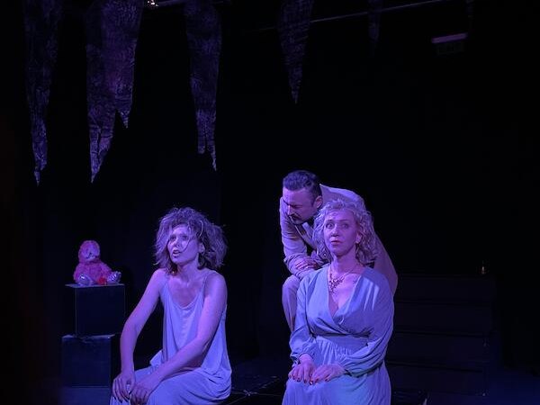 Drei Personen in einem dunklen Raum, dramatisch beleuchtet in Violett. Zwei sitzen auf Kisten, eine steht dahinter. Die Szene wirkt theatralisch., © Theater Atelier Drei Personen in einem dunklen Raum, dramatisch beleuchtet in Violett. Zwei sitzen auf Kisten, eine steht dahinter. Die Szene wirkt theatralisch., © Theater Atelier