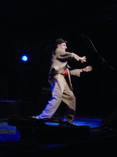 Ein Mann in einem Anzug und Hut gestikuliert lebhaft auf einer Bühne mit Mikrofon, umgeben von dunkler Beleuchtung., © Theaterhaus Stuttgart e.V.