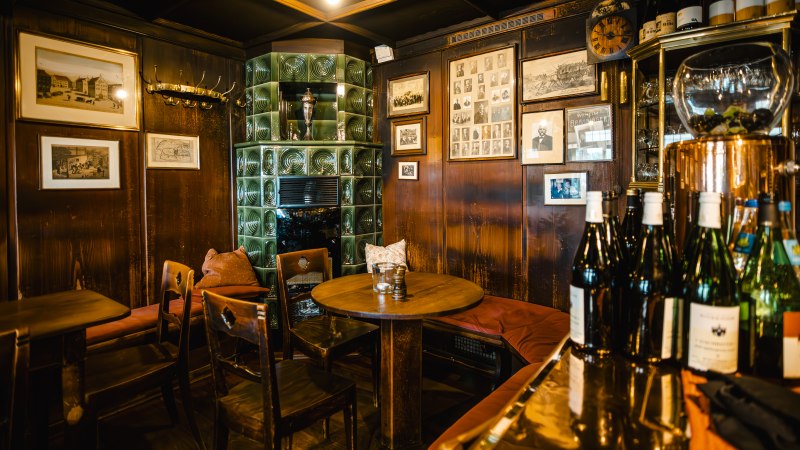 Gemütliche Weinstube mit Holzmöbeln, grünem Kachelofen, Bildern an den Wänden und Weinflaschen auf einem Tisch., © SMG Stuttgart Marketing GmbH - Sarah Schmid