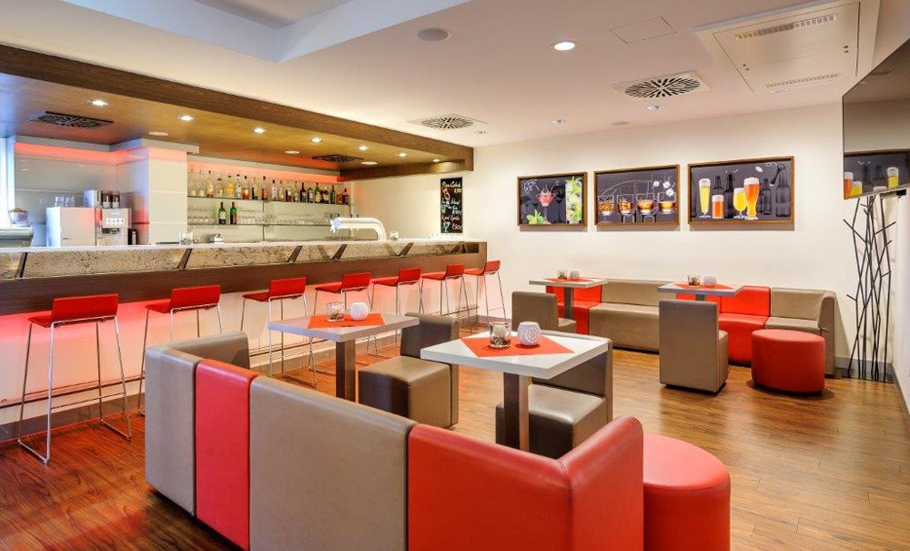 Moderne Bar mit roten und grauen Möbeln, beleuchtetem Tresen und Wandbildern von Getränken. Elegantes Ambiente mit Holzboden., © Ibis Stuttgart Airport Messe