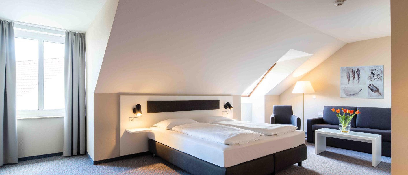 Bright hotel room with double bed, sofa, table and flowers. Sloping ceiling, window with curtains, modern furnishings., &copy; Erikson Hotel