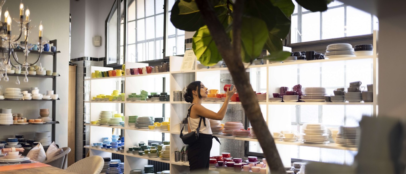 Frau in einem Geschäft betrachtet bunte Geschirrteile in Regalen. Der Raum ist hell erleuchtet, mit großen Fenstern und einem Kronleuchter., © SMG, Ingolf Pompe Frau in einem Geschäft betrachtet bunte Geschirrteile in Regalen. Der Raum ist hell erleuchtet, mit großen Fenstern und einem Kronleuchter., © SMG, Ingolf Pompe