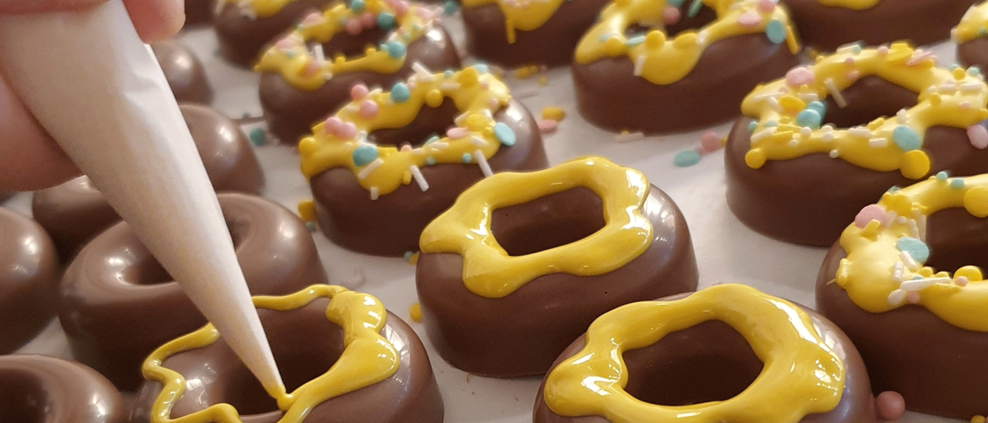 Chocolate rings are decorated with yellow icing and colorful sprinkles. One hand holds a piping bag., © kesselglueck.de