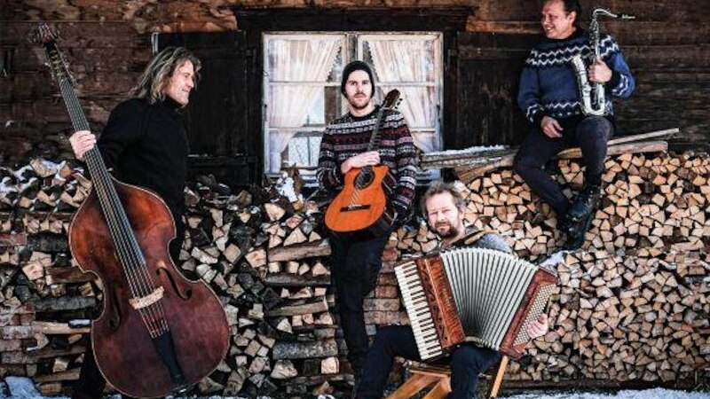 Vier Musiker posieren mit Kontrabass, Gitarre, Akkordeon und Saxophon vor einer Holzhütte auf einem Holzstapel im Schnee., © StuttgartKonzert Veranstaltungs GmbH Vier Musiker posieren mit Kontrabass, Gitarre, Akkordeon und Saxophon vor einer Holzhütte auf einem Holzstapel im Schnee., © StuttgartKonzert Veranstaltungs GmbH