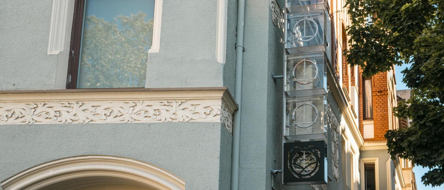 Fassade eines Gebäudes mit verzierten Elementen und einem Schild mit Logo. Ein Baum wirft Schatten auf die Wand. Klare, sonnige Atmosphäre., © SMG, Sarah Schmid