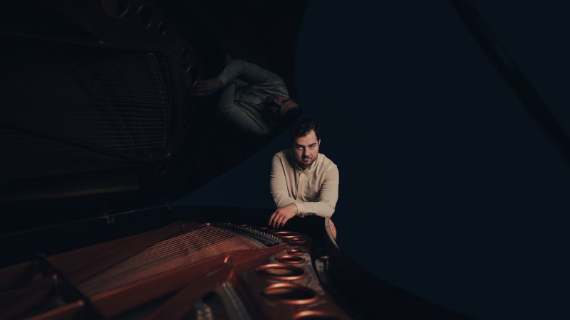 Ein Mann lehnt sich über einen Flügel, sein Spiegelbild ist im Deckel sichtbar. Der Hintergrund ist dunkel gehalten., © Clara Evens