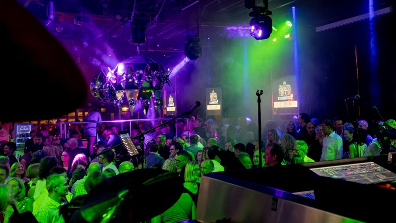 Eine lebhafte Party mit vielen Menschen, bunten Lichtern und einem DJ-Pult im Vordergrund. Im Hintergrund sind Bildschirme mit "Älternabend Silvesterparty" zu sehen., © Adventure Perkins GmbH Eine lebhafte Party mit vielen Menschen, bunten Lichtern und einem DJ-Pult im Vordergrund. Im Hintergrund sind Bildschirme mit "Älternabend Silvesterparty" zu sehen., © Adventure Perkins GmbH