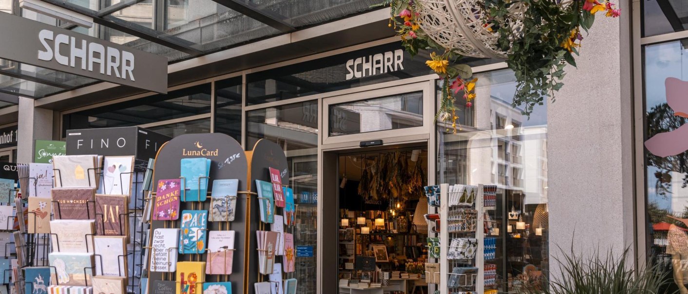 Eingang der Buchhandlung Scharr mit Kartenständern voller Grußkarten und hängenden Blumenarrangements. Innen sind Bücher und Lampen sichtbar., © Stuttgart Marketing GmbH, Sarah Schmid