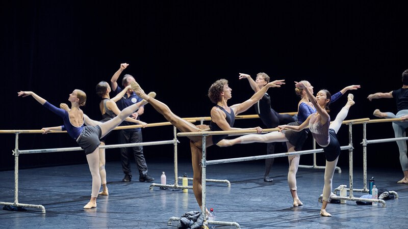 Ballettt&auml;nzer &uuml;ben an der Ballettstange auf einer B&uuml;hne. Sie tragen Trainingskleidung und f&uuml;hren Bein&uuml;bungen aus., &copy; W&uuml;rttembergische Staatstheater Stuttgart