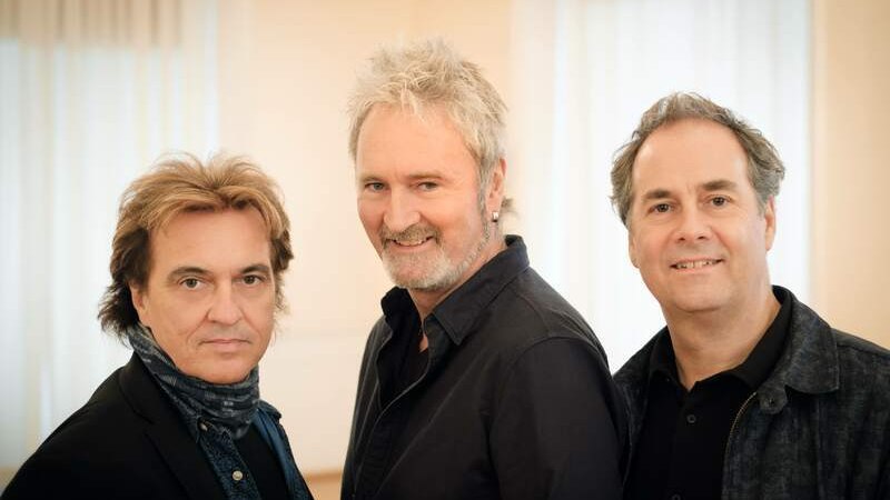 Drei M&auml;nner stehen nebeneinander und l&auml;cheln in die Kamera. Sie tragen dunkle Kleidung und stehen vor einem hellen, unscharfen Hintergrund., &copy; C2 Concerts GmbH