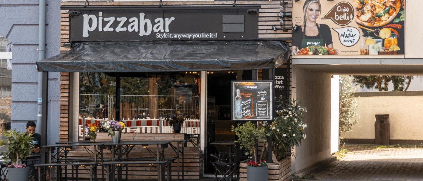 Außenansicht einer Pizzabar mit Terrasse, Blumen und einem Werbeplakat. Ein Mann sitzt an einem Tisch. Die Markise ist schwarz., © Stuttgart Marketing GmbH, Sarah Schmid