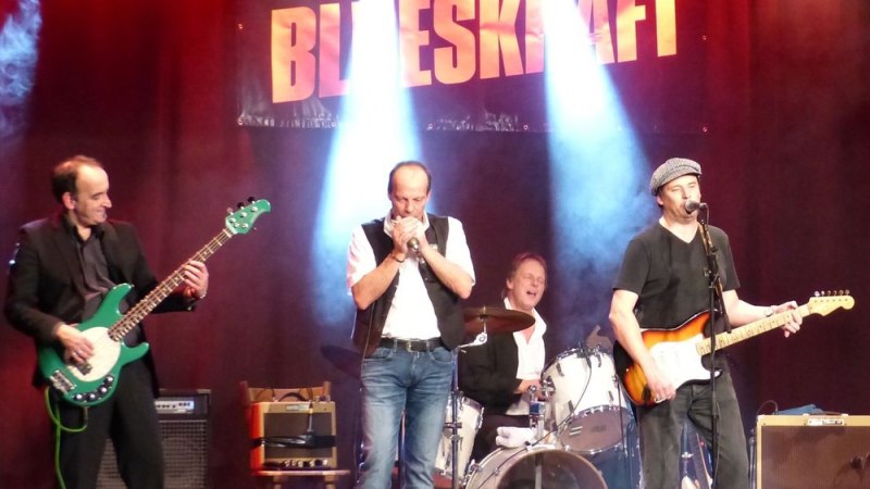 Eine Bluesband performt auf der B&uuml;hne. Der Bassist spielt eine gr&uuml;ne Gitarre, der S&auml;nger spielt Mundharmonika, der Schlagzeuger sitzt im Hintergrund und der Gitarrist singt., &copy; Veranstalter