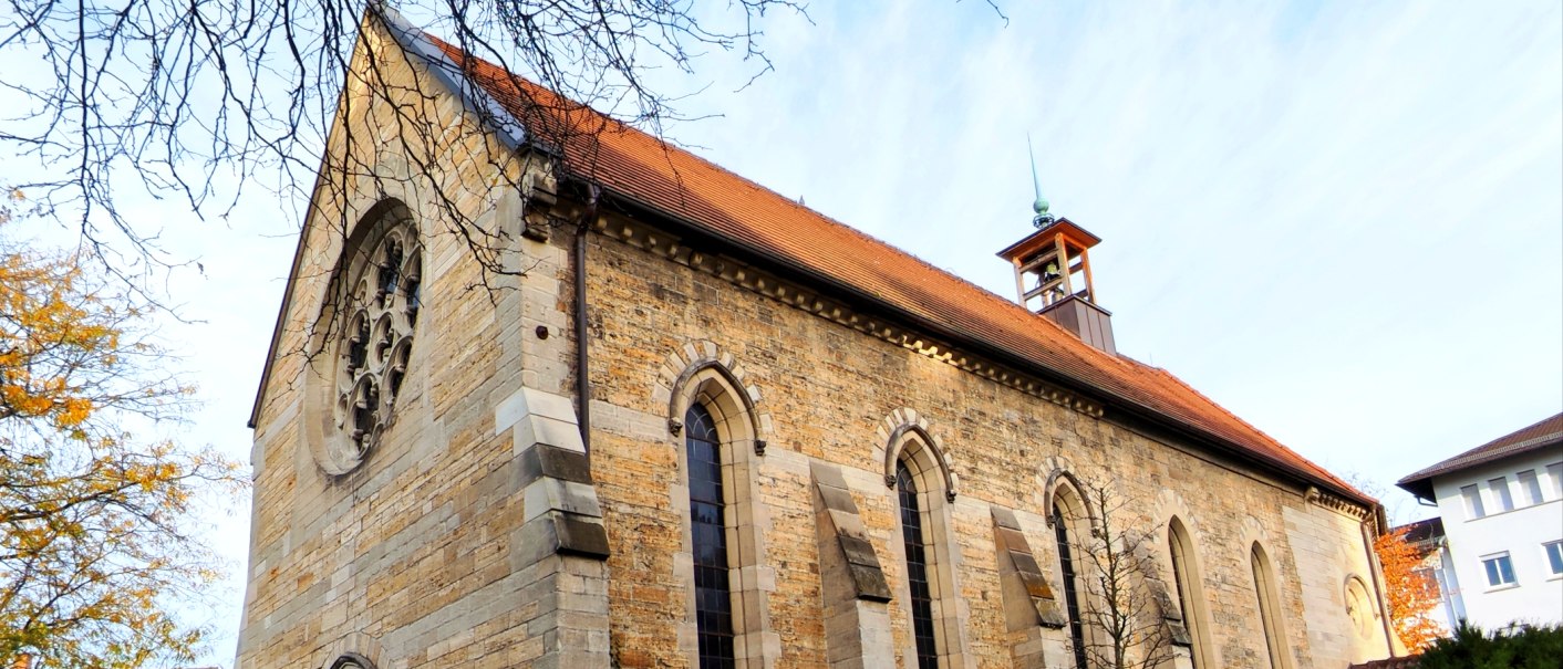 Steinkirche mit rotem Satteldach, Rundfenster und kleinem Glockenturm, von kahlen Ästen und Herbstbäumen umrahmt., © St. Katharina Stuttgart Steinkirche mit rotem Satteldach, Rundfenster und kleinem Glockenturm, von kahlen Ästen und Herbstbäumen umrahmt., © St. Katharina Stuttgart