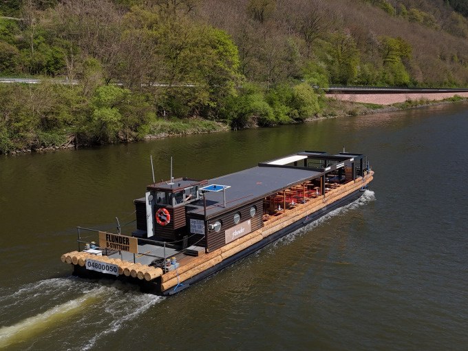 Das Flussschiff Flunder f&auml;hrt auf einem ruhigen Fluss, umgeben von gr&uuml;ner Landschaft. An Bord sind Tische und St&uuml;hle f&uuml;r Passagiere., &copy; Cool-Tours StattReisen