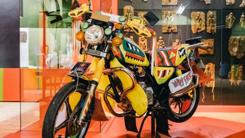 Ein farbenfrohes Motorrad mit kunstvollen Mustern steht in einem Museum. Im Hintergrund sind afrikanische Skulpturen und Reliefs zu sehen., © Linden-Museum Stuttgart