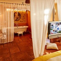 Ein gemütliches Zimmer mit Badewanne, Fernseher und weißen Vorhängen. Der Boden ist aus Terrakottafliesen, ein Sessel steht in der Ecke., © Designhotel & Restaurant Der Zauberlehrling